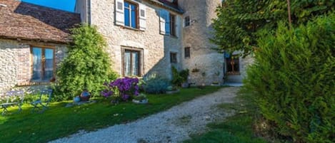 A stone house with a round tower, a gravel path, and a garden with flowers and shrubs.