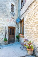 Un estrecho patio de piedra con una puerta de madera, una ventana con contraventanas de madera y plantas en macetas.