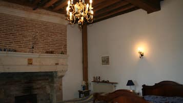 A traditional room with a brick fireplace, a wooden bed with a patterned bedspread, a chandelier, and a wall-mounted lamp.