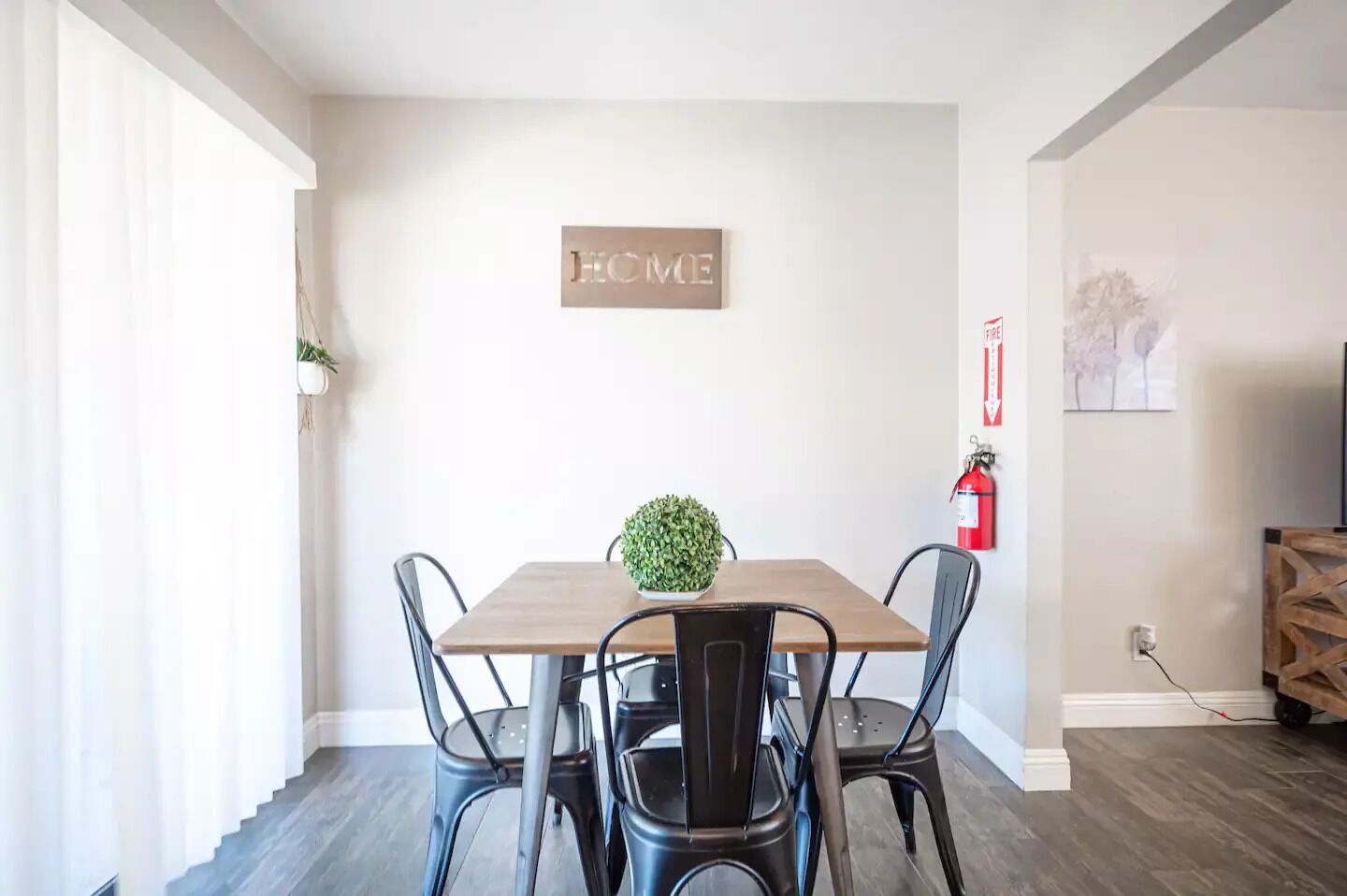 Un comedor con mesa de madera, cuatro sillas, una planta pequeña en maceta, un cartel con la palabra 'HOME', un extintor y una toma de corriente.