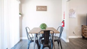 A dining area with a wooden table, four chairs, a small potted plant, a 'HOME' sign on the wall, a fire extinguisher, and a power outlet.