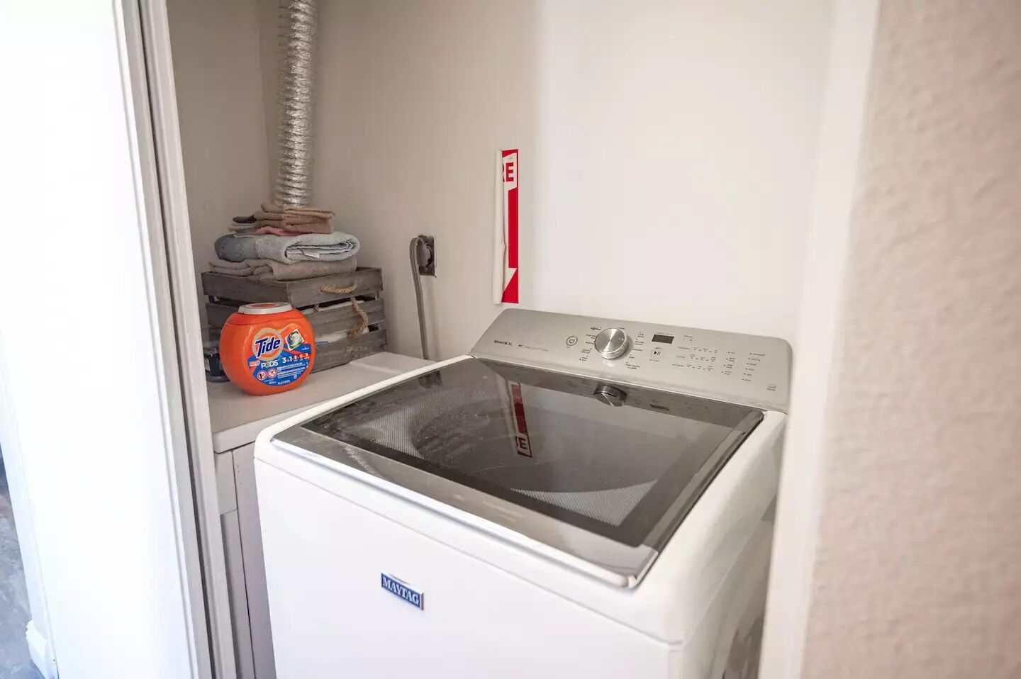 Etukuormattava Maytag-pesukone ja -kuivausrumpu pyykinhuoneessa, jossa on myös Tide-pesuainetta ja punainen palosammutin seinällä.