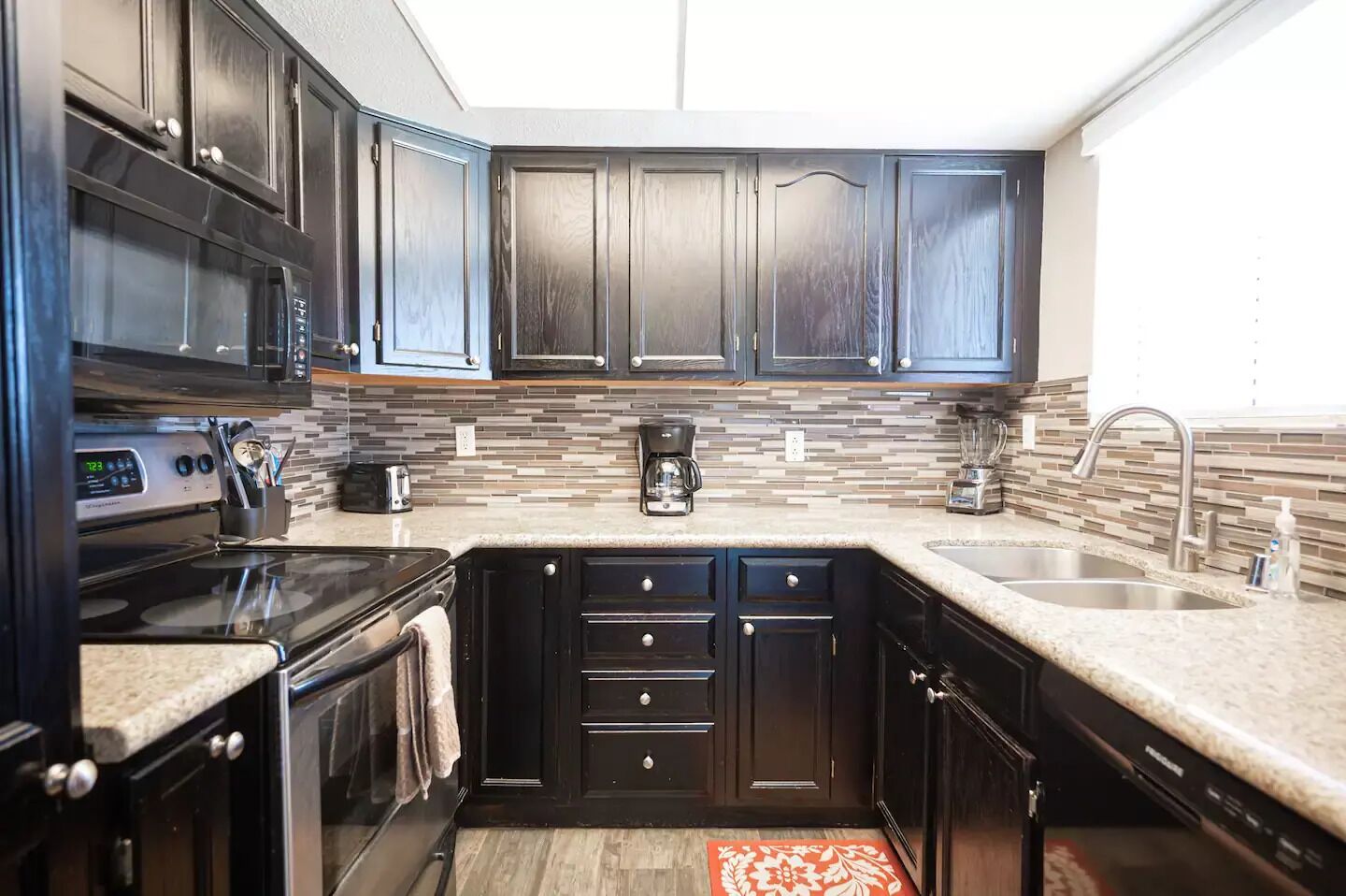 A modern kitchen with dark wood cabinets, a granite countertop, a built-in oven, a coffee maker, and a dishwasher.