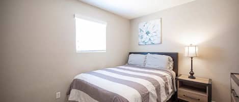 Een slaapkamer met een bed, nachtkastje, dressoir, raam en plafondventilator.