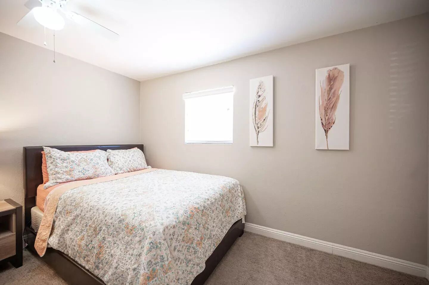 Un dormitorio con cama, mesita de noche, ventilador de techo y dos ventanas.
