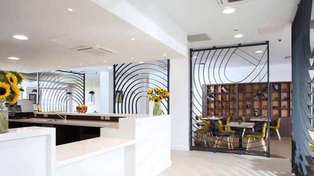 A modern reception area with a white counter, a glass partition with a sunflower decoration, and a seating area with yellow chairs and a wooden bookshelf. - Sunley Hotel (Northampton)