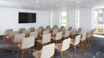 A conference room with rows of grey chairs and orange frames, a flat-screen TV on the wall, and large windows offering a view of the outside.
