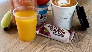 Une table avec une bouteille d’eau, une tasse de café, une banane, un jus de fruits, un paquet de chips, un paquet de pain et un paquet de barres de céréales.