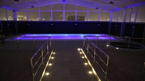 An indoor swimming pool with a view of the city at night, surrounded by railings and illuminated by ceiling lights. - Quetzal (Riyadh)