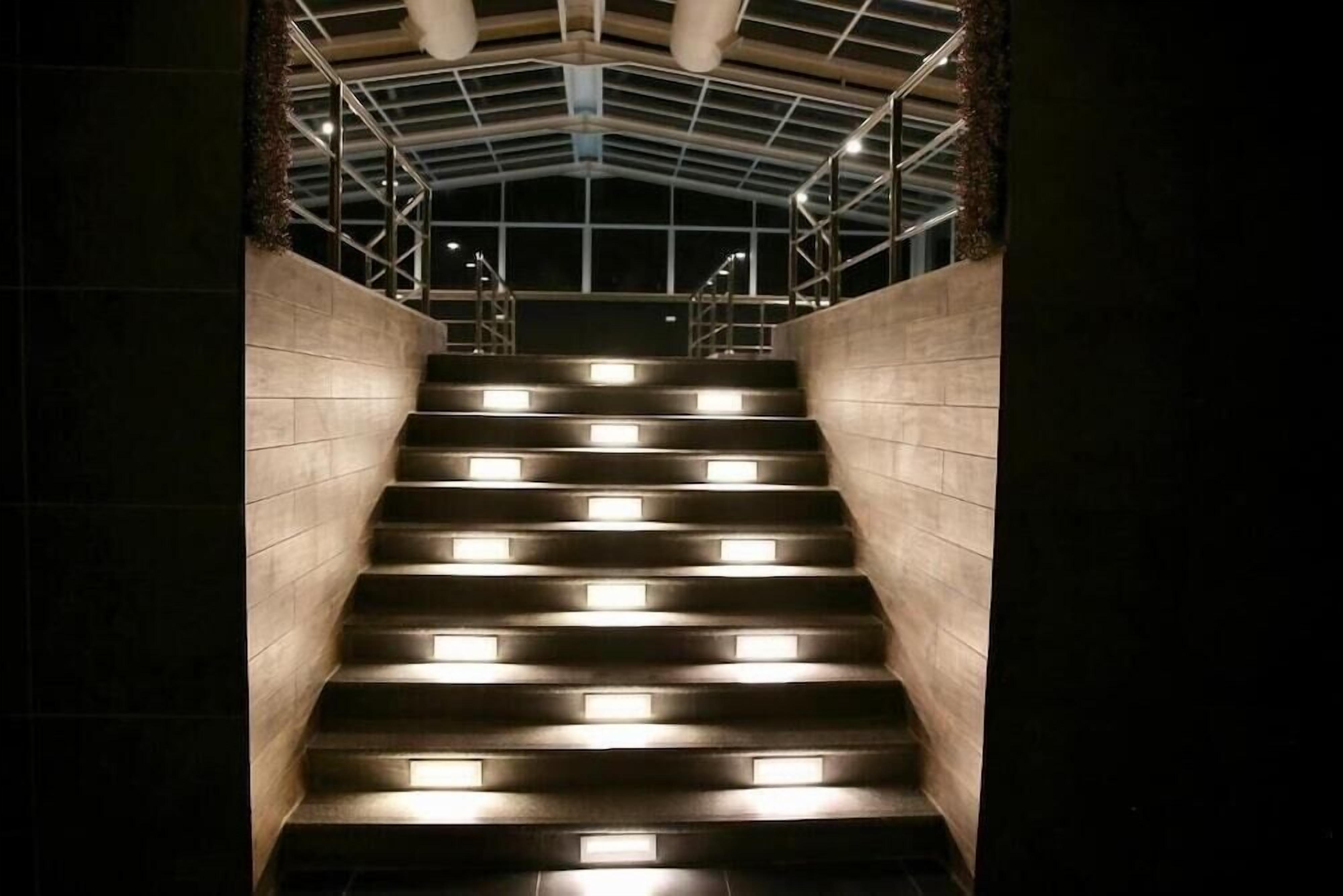 A staircase with illuminated steps leading upwards, surrounded by walls and a metal framework ceiling.