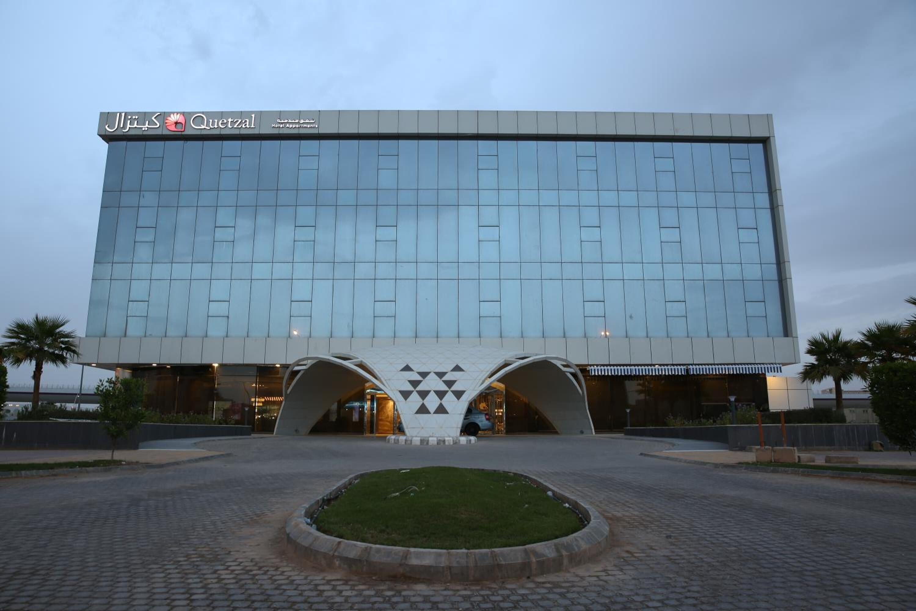 Un hotel moderno con fachada de vidrio reflectante, una entrada central con diseño geométrico y un patio empedrado con un pequeño jardín.