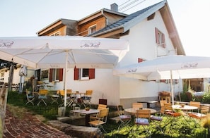 An outdoor seating area with tables and chairs, umbrellas, and a building with a sloped roof in the background.