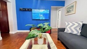 Una sala de estar con una pared azul decorativa, un televisor montado sobre una planta, un sofá con cojines estampados y piso de madera.