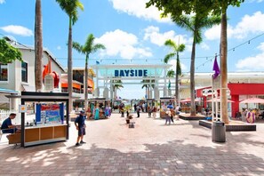 Uma área de compras ao ar livre movimentada, com uma placa grande que diz 'BAYSIDE', palmeiras e diversas lojas e restaurantes.