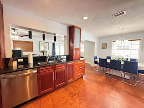 A kitchen with a stainless steel dishwasher, wooden cabinets, a black countertop, and a dining area with blue chairs and a chandelier.