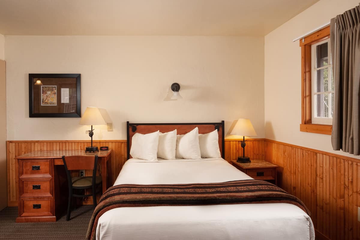 Une chambre d’hôtel avec un lit en bois, un bureau et une chaise. Il y a une fenêtre avec des rideaux, une lampe fixée au mur et un tableau encadré accroché au mur.
