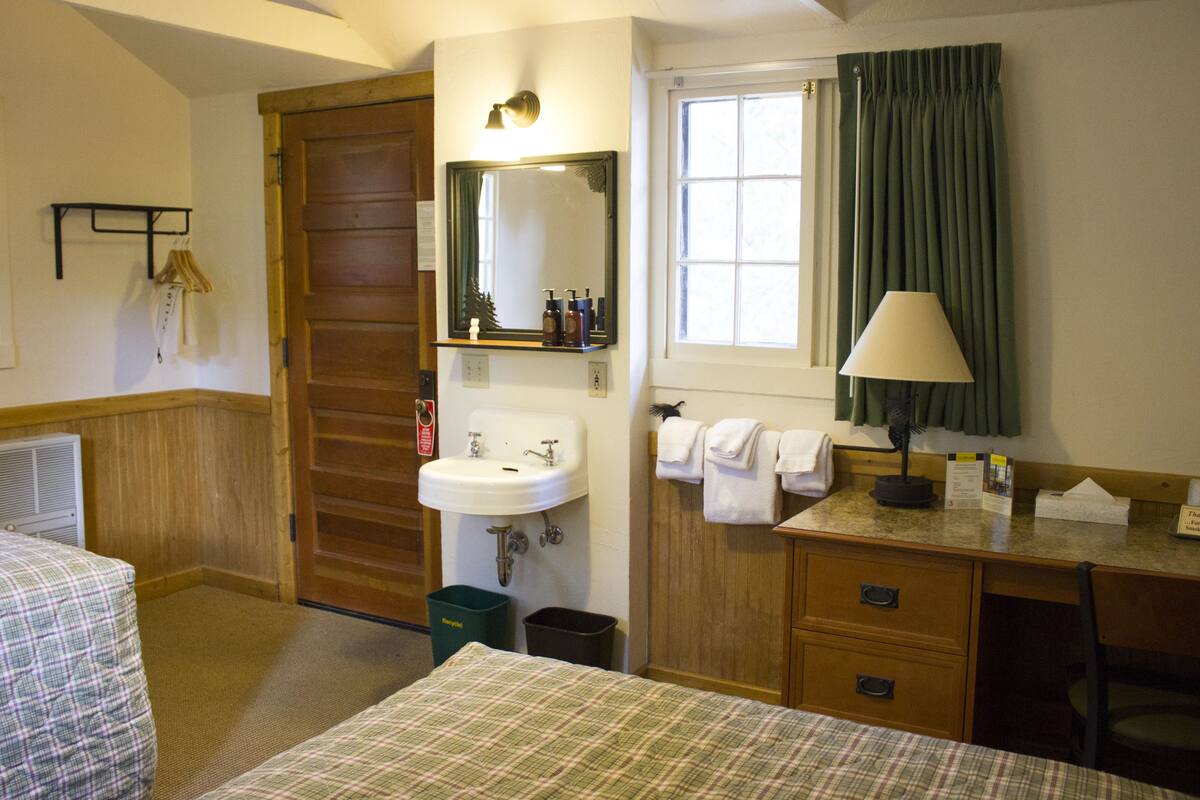 Une chambre d’hôtel avec un lit, une porte en bois, un lavabo, un miroir, une fenêtre avec des rideaux et un bureau avec une lampe.