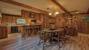 A rustic kitchen with wooden cabinets, a dining area with a large wooden table and bar stools, and a wooden floor. - Perfect location, creekside, close to skiing, easy paved access, Game Pavilion! (Maggie Valley)