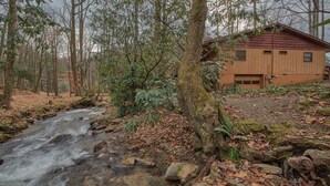 A cabin with a wooden exterior, surrounded by a forest and a flowing stream. - Perfect location, creekside, close to skiing, easy paved access, Game Pavilion! (Maggie Valley)