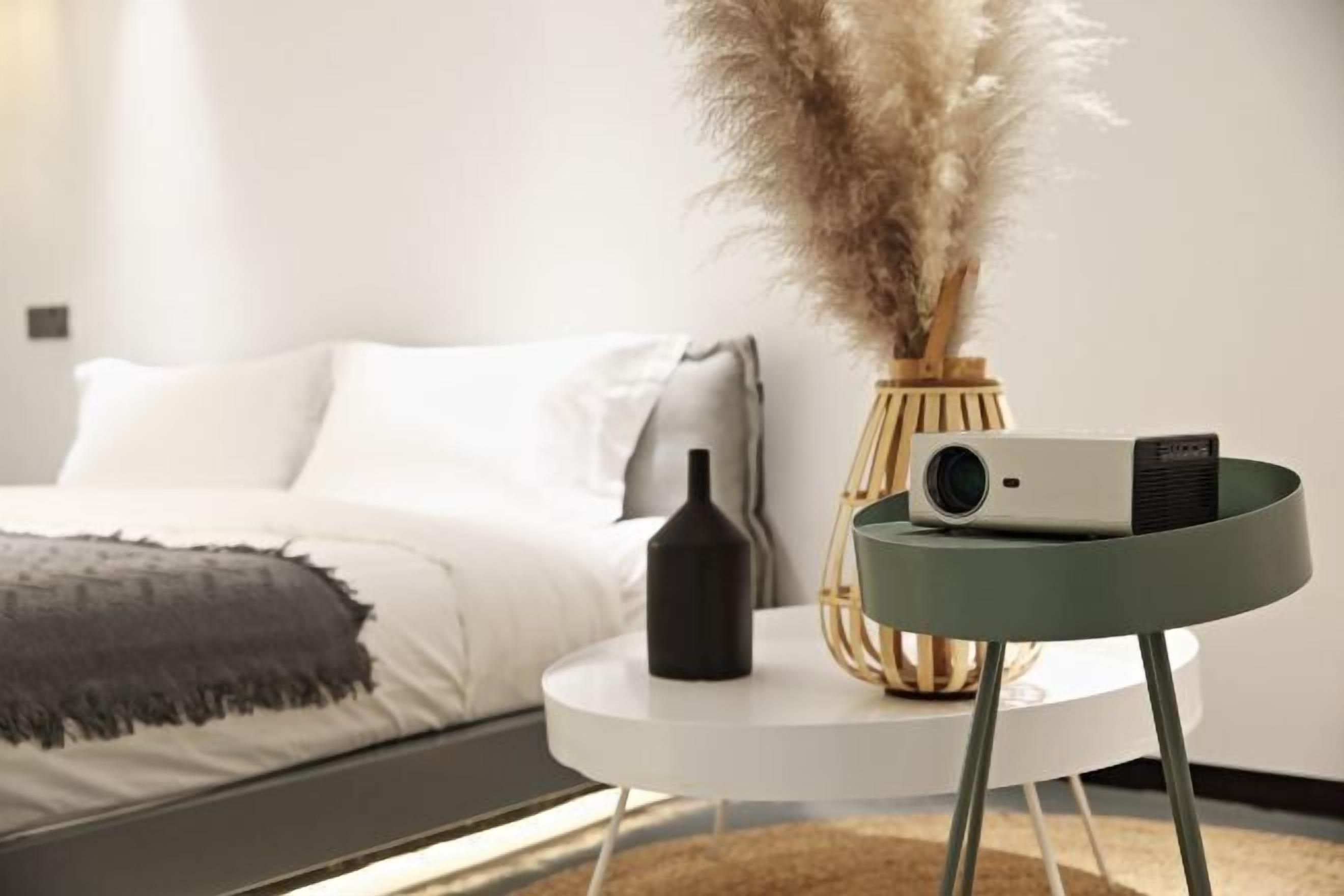 Un dormitorio ordenado con una cama, una mesita redonda con un proyector, un florero con plumas de pampa y una botella.