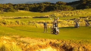 A golf course with sand bunkers, a putting green, and a distant tree line.