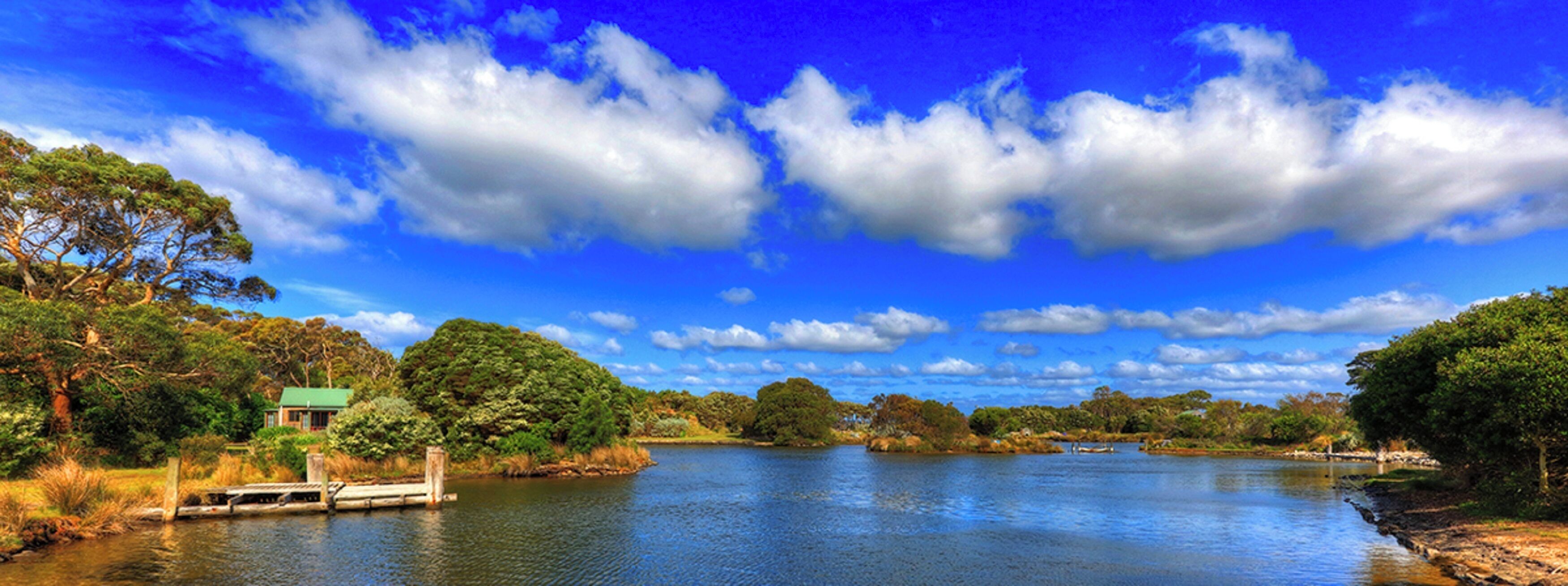 A serene riverside landscape with a wooden dock, lush greenery, and a clear blue sky.