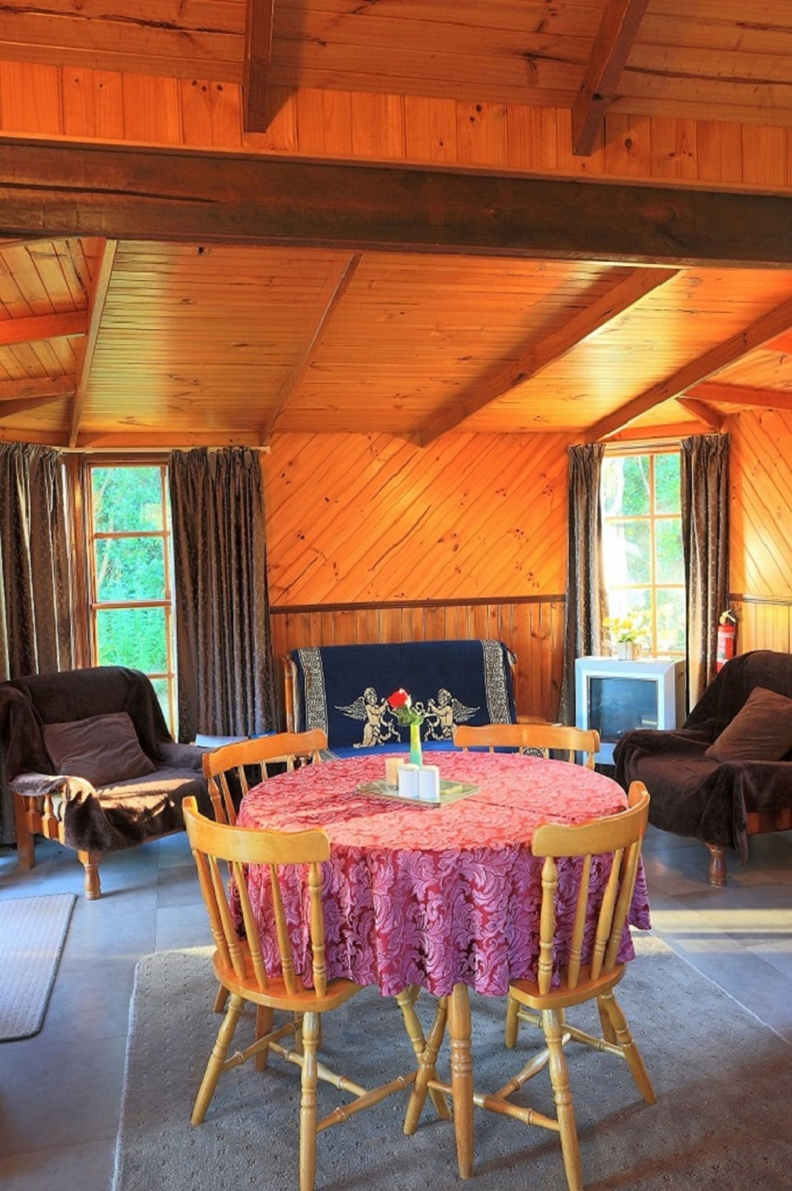 Een knus vertrek met een houten plafond, een ronde tafel met stoelen, een televisie en een raam met gordijnen.