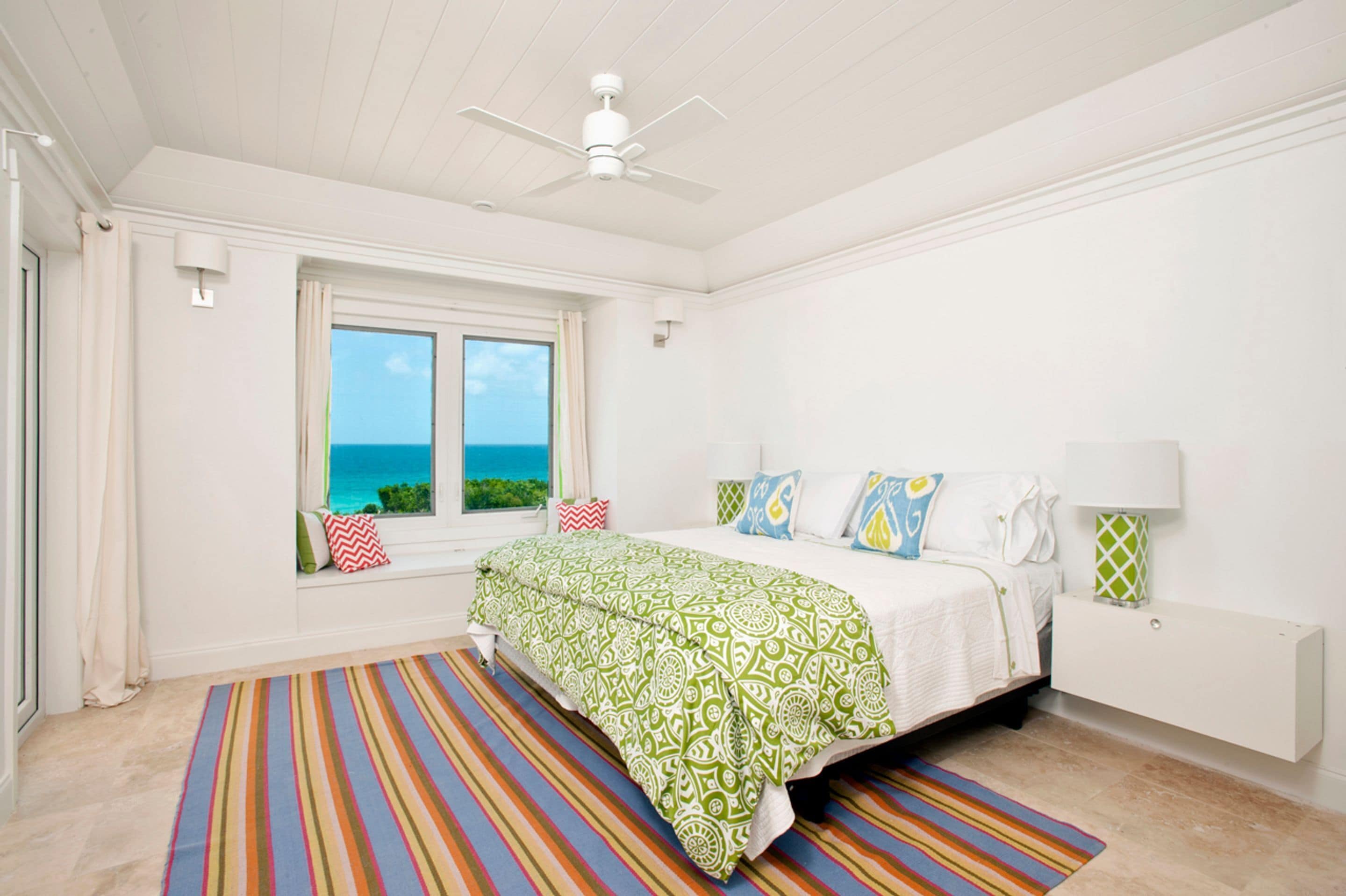 Een slaapkamer met een bed, een raam met uitzicht op de oceaan, een plafondventilator en een kleurrijk gestreept tapijt.