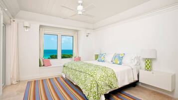 A bedroom with a bed, a window with a view of the ocean, a ceiling fan, and a colorful striped rug.