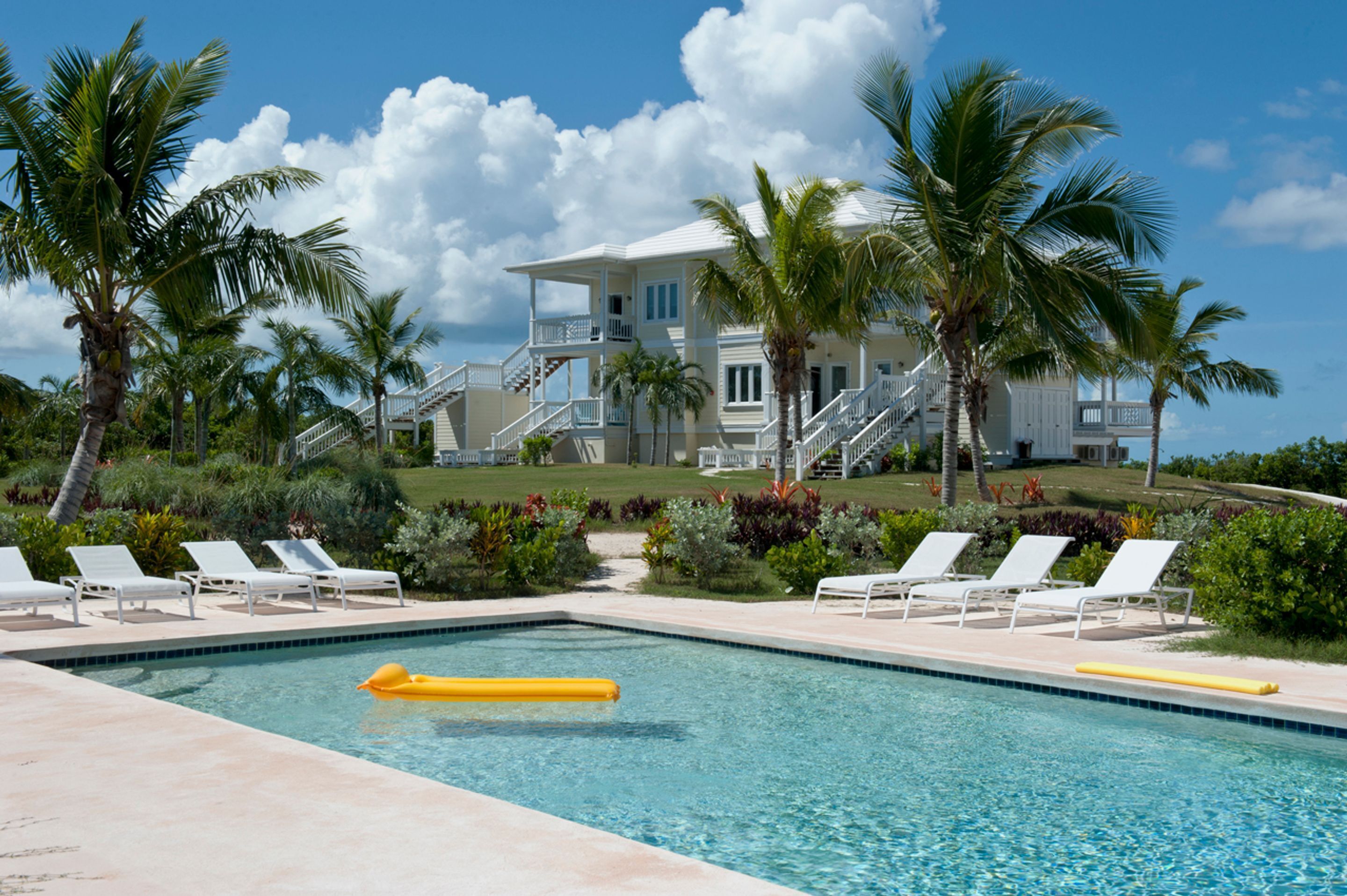 A pool with lounge chairs and a yellow float, surrounded by palm trees and a large building with a staircase.