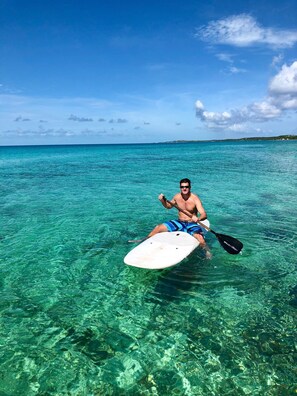 A man sitting on a paddleboard in clear turquoise water, with a distant shoreline and a clear sky. - Buttonwood Reserve 2A (James Cistern)