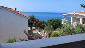 A coastal view with a clear sky, a building with a red-tiled roof, and a balcony overlooking the sea.