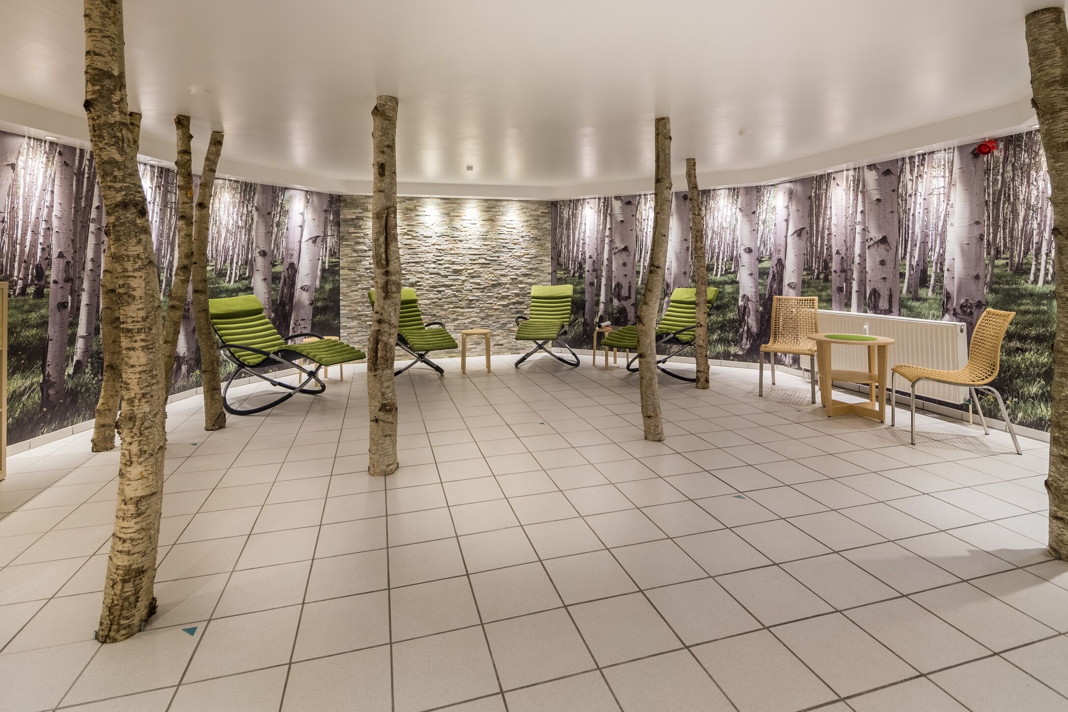 Ein Zimmer mit Birkenholzsäulen, grünen Lounge-Stühlen, einem Holztisch und einer Steinwand.