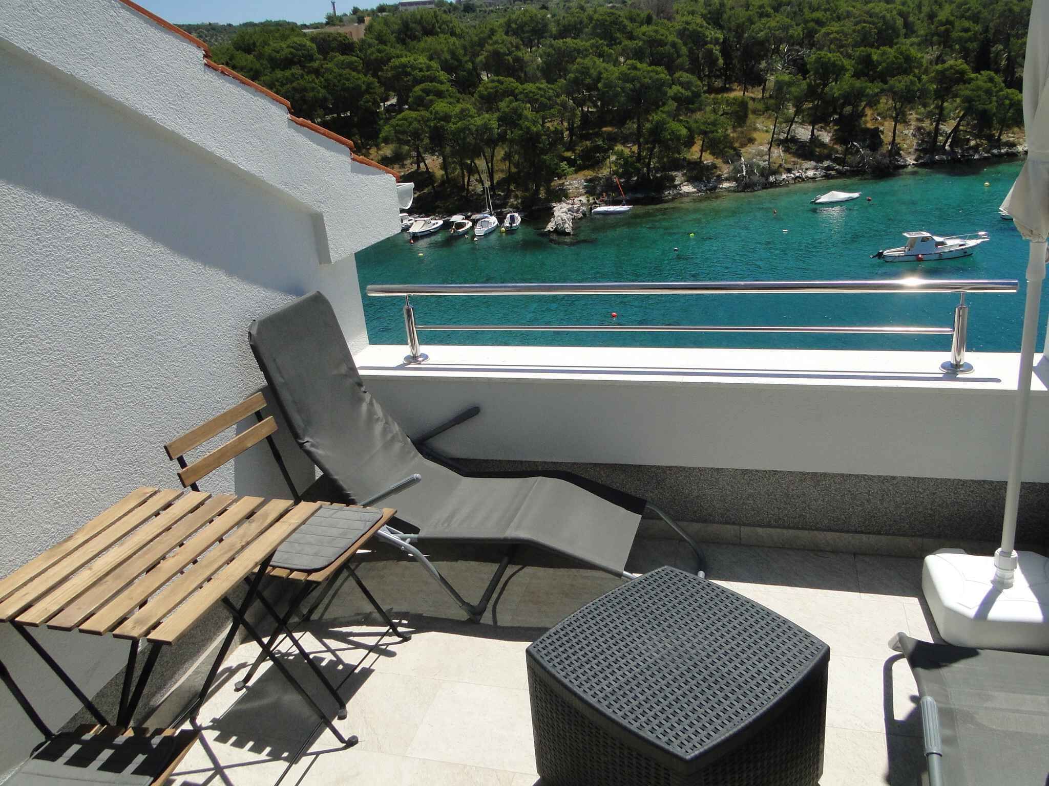 A balcony with a wooden table and two chairs, overlooking a turquoise sea with boats and a forested hillside.