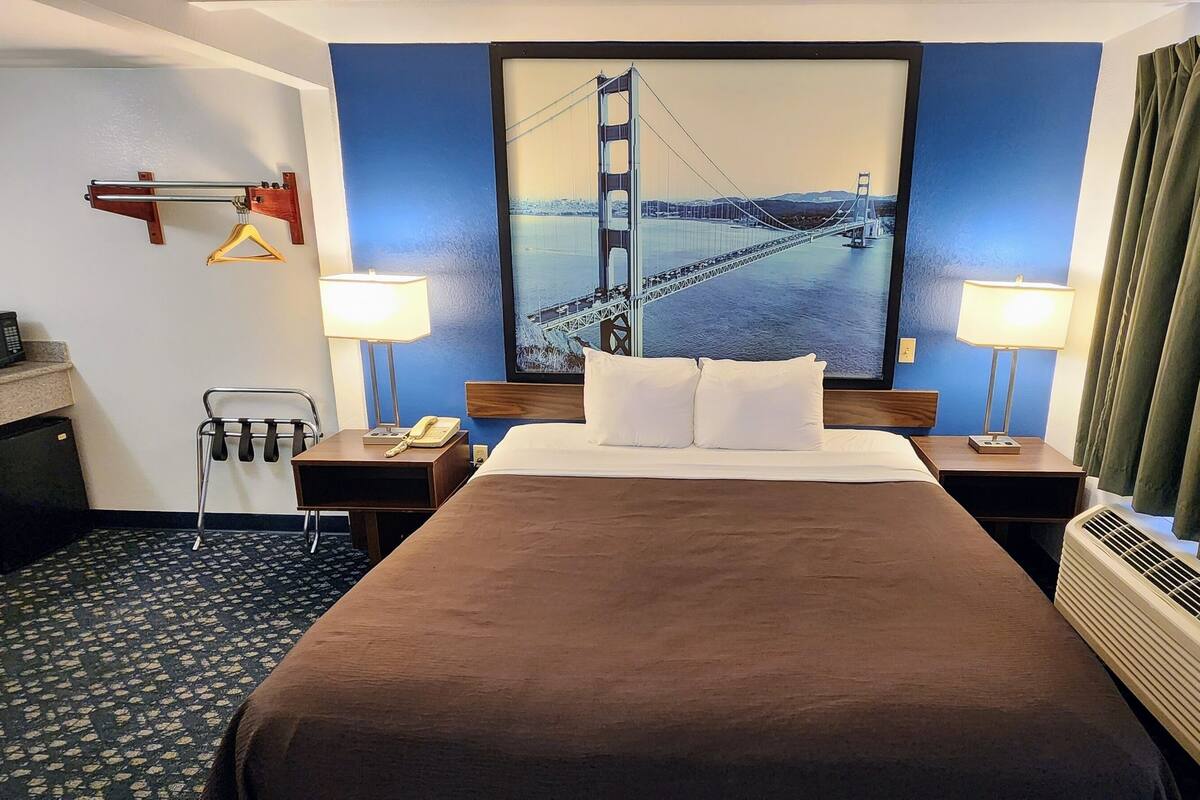A hotel room with a large bed, two bedside tables, a desk with a microwave, a hanging coat rack, and a painting of the Golden Gate Bridge.