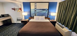 A hotel room with a large bed, two bedside tables, a desk with a microwave, a hanging coat rack, and a painting of the Golden Gate Bridge. - Hotel Belmont (Belmont)