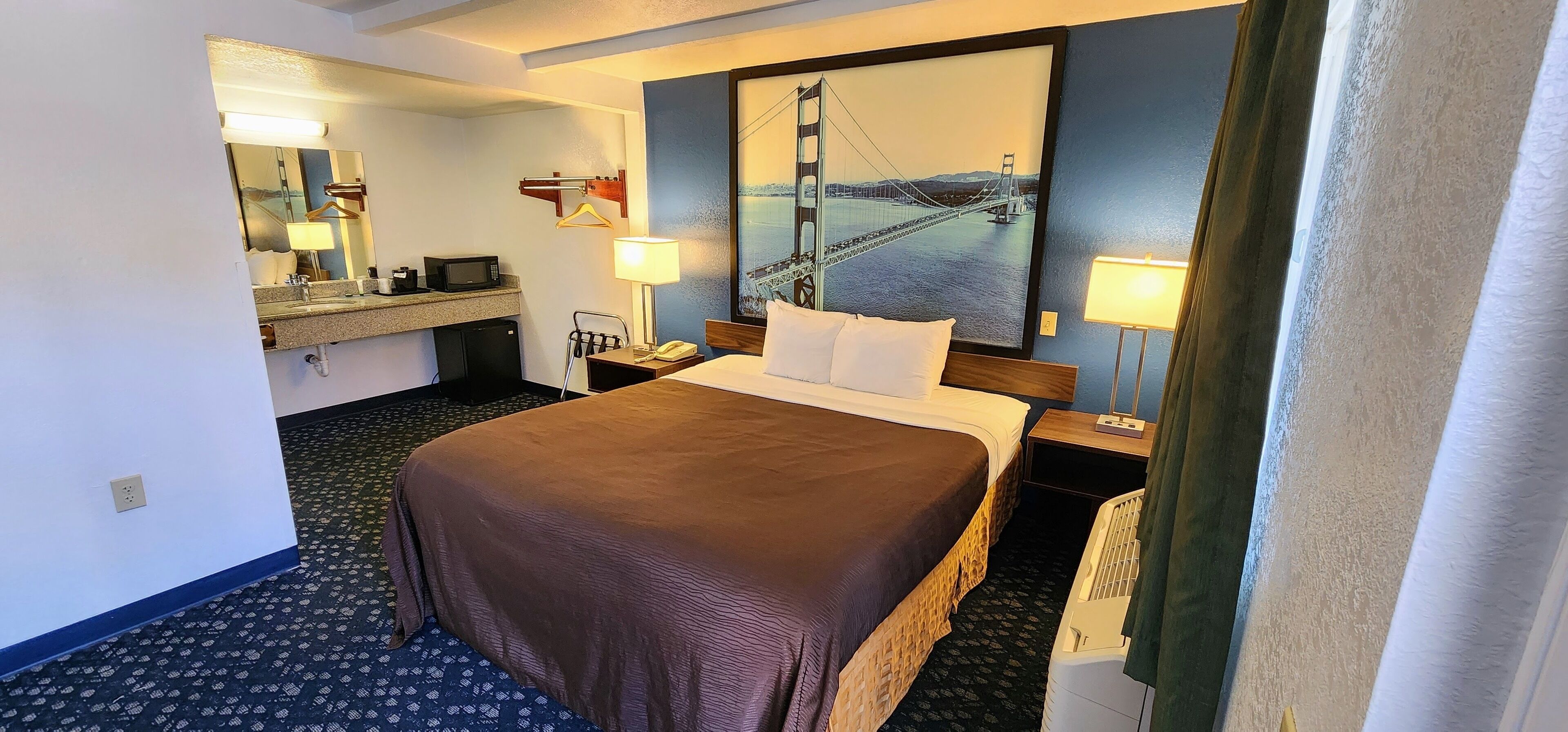Quarto de hotel com uma cama grande, uma escrivaninha com computador, um espelho, um cabideiro e um quadro de uma ponte.