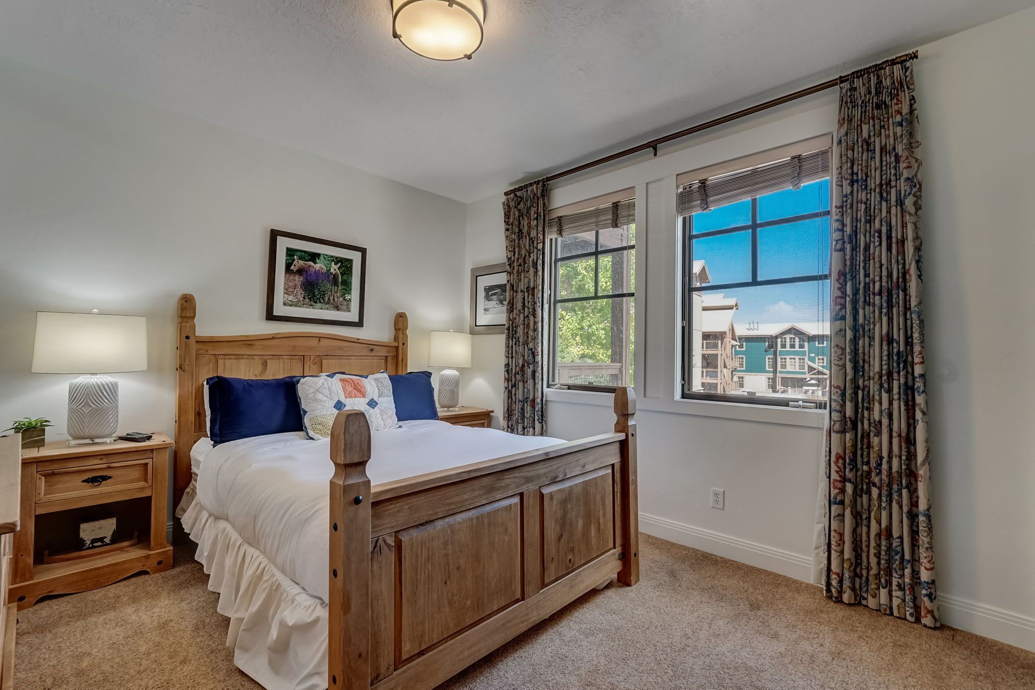 A bedroom with a wooden bed, a nightstand with a lamp, a window with curtains, and a view of buildings outside.
