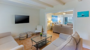 Ein Wohnzimmer mit Flachbildfernseher, zwei Sofas, einem Glastisch und einem Sessel. An der Wand hängt ein Gemälde und ein Fenster ist mit Jalousien versehen.