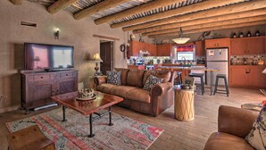 A rustic living room with a leather sofa, wooden coffee table, and a television on a cabinet. A kitchen is visible in the background with wooden cabinets and a bar stool. - El Prado Adobe Home: Courtyard w/ Mountain Views! (El Prado)