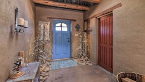 A rustic hallway with a blue door, wooden beams, a wall-mounted light fixture, and a small table with a decorative item. - El Prado Adobe Home: Courtyard w/ Mountain Views! (El Prado)