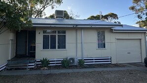 A single-story house with a metal roof, a front door, two windows, a garage door, and a small garden area with plants.