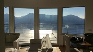 A room with a large window overlooking a mountainous landscape and a body of water. There is a sofa and a table with bags on it. - Superb villa with panoramic view over Lake Maggiore (Pollino)