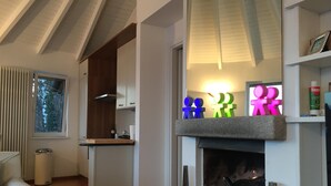 A modern living room with a fireplace, wooden beams on the ceiling, and a kitchen area with a window. - Superb villa with panoramic view over Lake Maggiore (Pollino)
