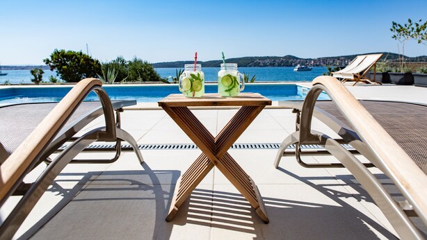 A poolside area with a table and two drinks, overlooking a body of water and hills. - EXCLUSIVE LOCATION BEACH APARTMENT "JAN"! NEW OPEN  SWIMMING POOL! (Trogir)