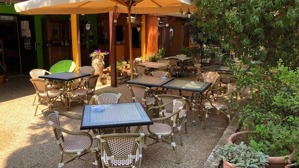 An outdoor patio with wicker furniture and a large umbrella, surrounded by potted plants and a building with a green facade.