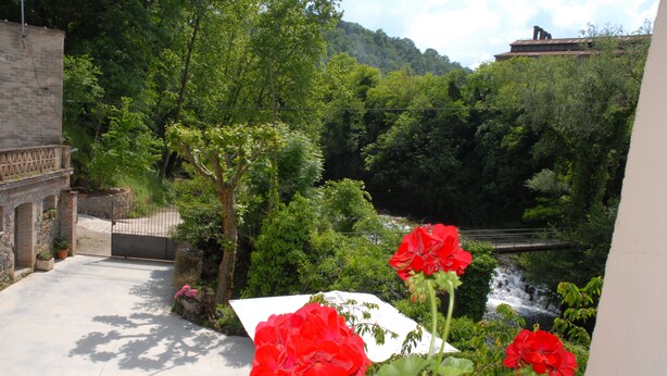 A courtyard with a paved walkway, red flowers, and a view of a waterfall and forested hillside. - CANXAI THE RURAL TOURIST HOUSE OF LA GARROTXA PER ENJOY AMB QUI MÉS ESTIMES (La vall de Bianya)