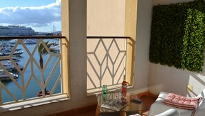 Ein Balkon mit Blick auf Boote, ein Glastisch mit einer Zeitschrift und ein weißer Stuhl mit gestreifter Sitzkissen.