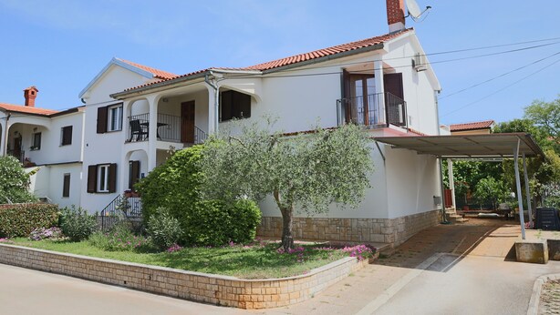 Une maison à deux étages, avec un toit recouvert de tuiles rouges, une terrasse couverte et un jardin agrémenté de plantes à fleurs.