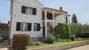 Une maison à deux étages avec un balcon, une porte d’entrée et un petit jardin agrémenté de fleurs et d’arbustes.